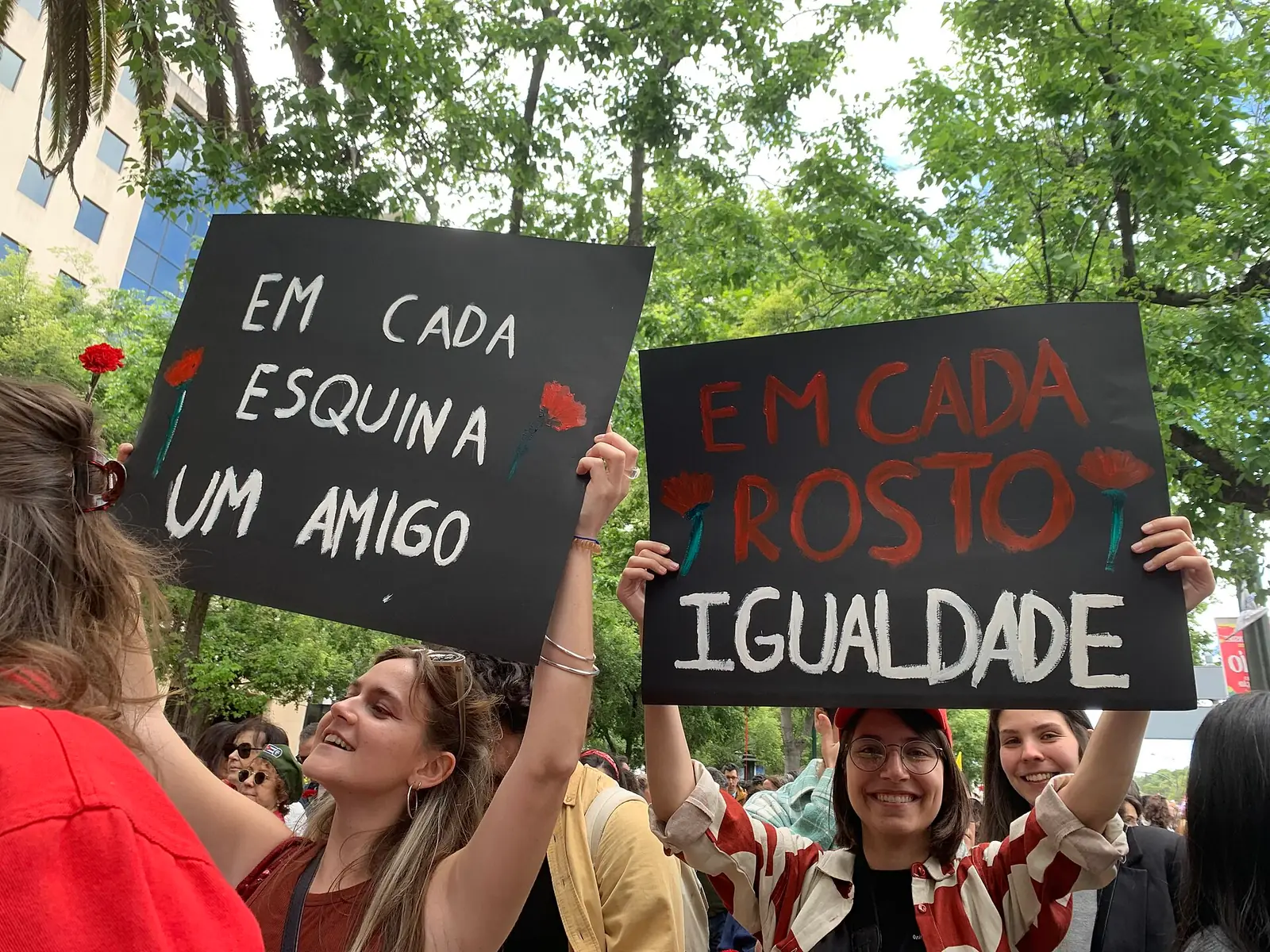 Duas jovens sorridentes levantam cartazes pretos escritos à mão, com cravos vermelhos desenhados, debaixo das árvores e no meio da multidão.