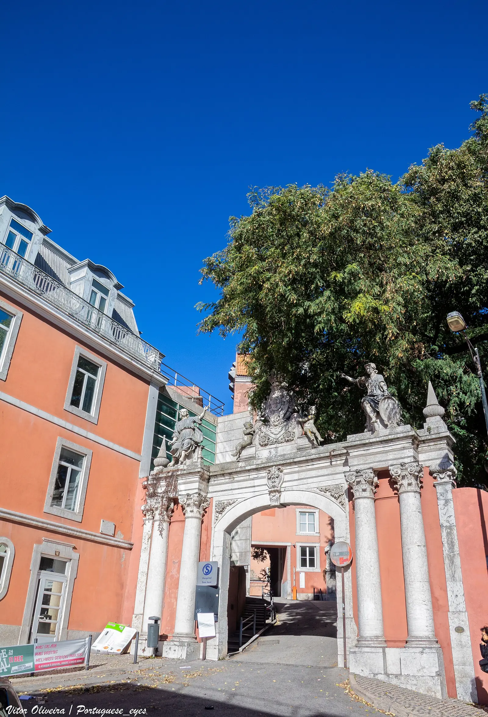 Portail de pierre claire en arc, couronné de sculptures, entre des murs couleur saumon ; à l’intérieur, une cour plantée d’arbres.
