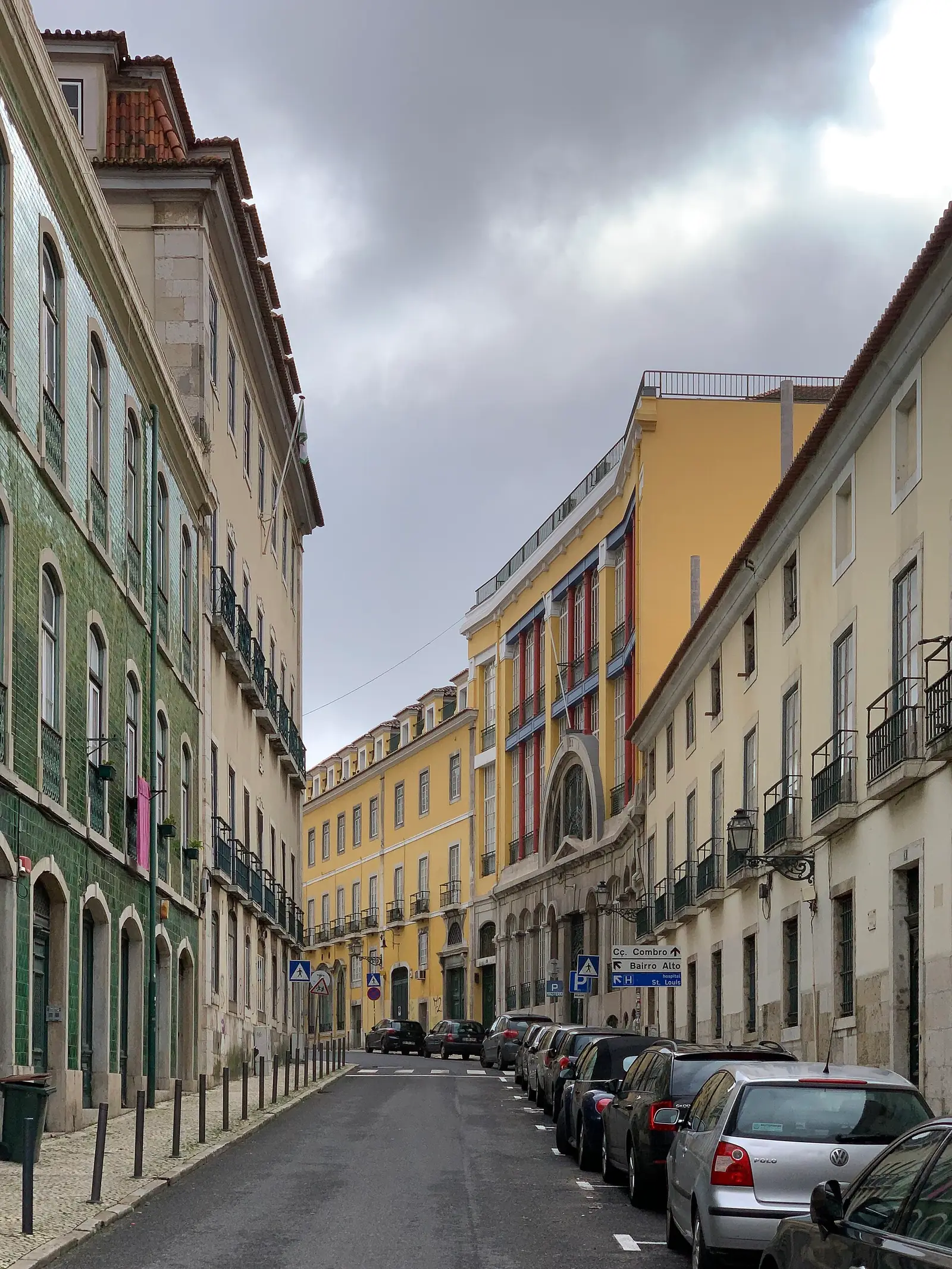 Rua comprida e ligeiramente inclinada entre prédios de três e quatro andares pintados de amarelo, verde e branco, com carros estacionados de um dos lados, sob céu nublado.