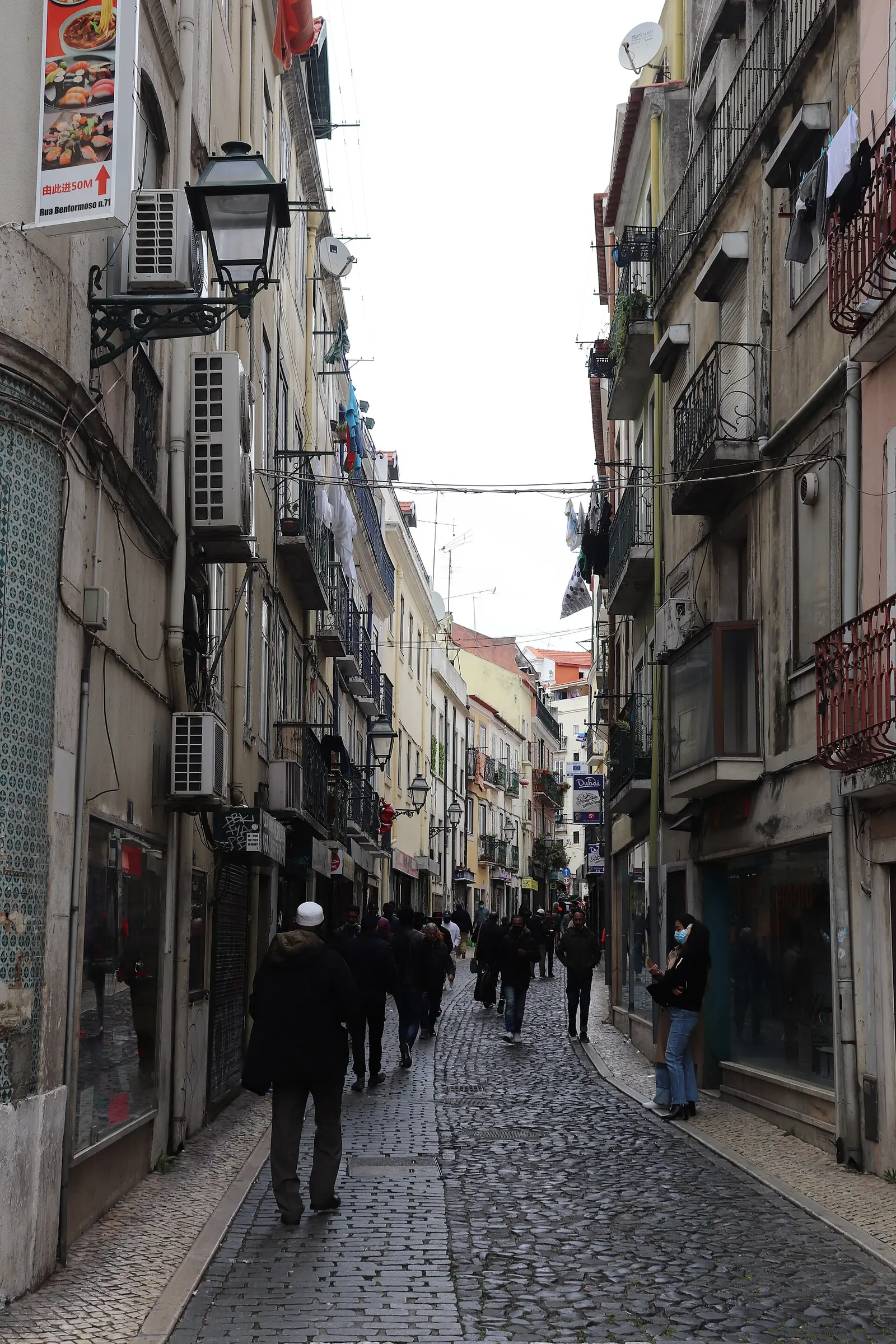 Calle estrecha y larga entre edificios altos con balcones, con tiendas en los bajos, ropa tendida y gente que pasa al fondo.
