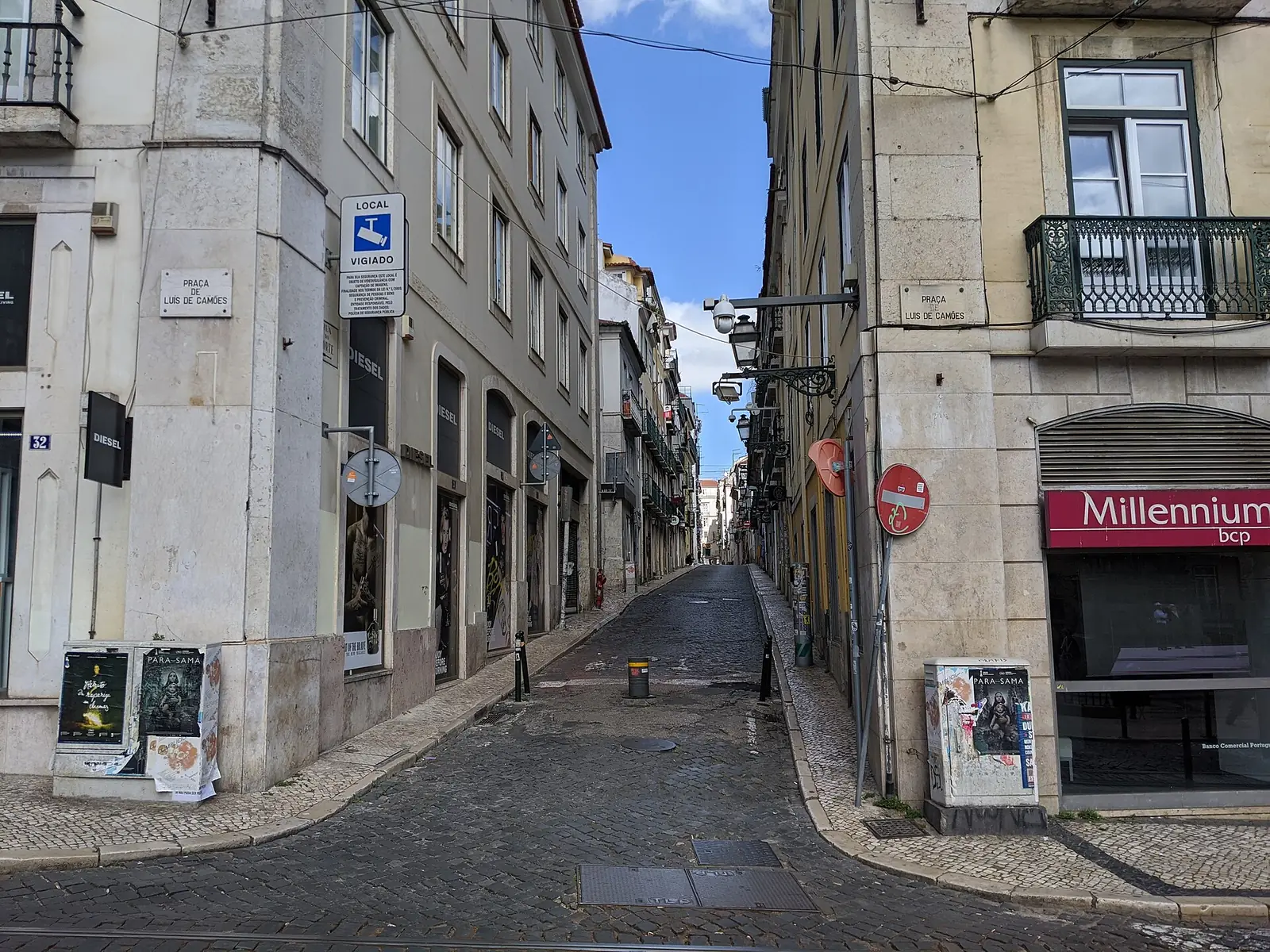 Rua estreita entre prédios claros de quatro andares, vista de uma esquina, com uma agência bancária no rés-do-chão à direita.